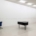 Installationsansicht „Œuvre (working title)“ von Sanna Helena Berger im Westfälischen Kunstverein: Ein schwarzer Tisch und ein blauer Bürostuhl stehen in einem hellen Ausstellungsraum. An den weißen Wänden hängen zahlreiche kleinformatige Blätter in einer langen Reihe.