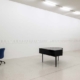 Installationsansicht „Œuvre (working title)“ von Sanna Helena Berger im Westfälischen Kunstverein: Ein schwarzer Tisch und ein blauer Bürostuhl stehen in einem hellen Ausstellungsraum. An den weißen Wänden hängen zahlreiche kleinformatige Blätter in einer langen Reihe.