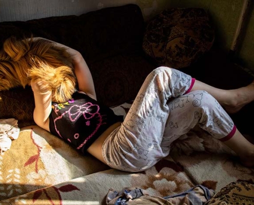 06_Versluis Fotografie „Nastya und Mila“ von Karine Zenja Versluis mit einem jungen Mädchen und einem Hund, die in einem von Sonnenlicht durchzogenen Innenraum auf einem Sofa liegen