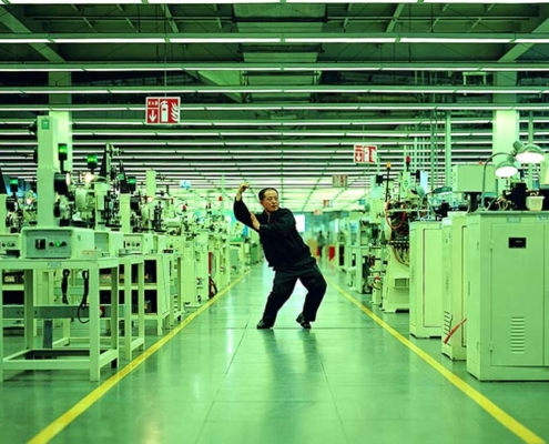 My_Future_is_Not_a_Dream Filmstill aus „Whose Utopia“ von Cao Fei aus dem Jahr 2006: Ein Mann nimmt eine tänzerische Pose in einer langen, grün beleuchteten Fabrikhalle zwischen Maschinen und Produktionsanlagen ein.