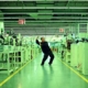 Filmstill aus „Whose Utopia“ von Cao Fei aus dem Jahr 2006: Ein Mann nimmt eine tänzerische Pose in einer langen, grün beleuchteten Fabrikhalle zwischen Maschinen und Produktionsanlagen ein.