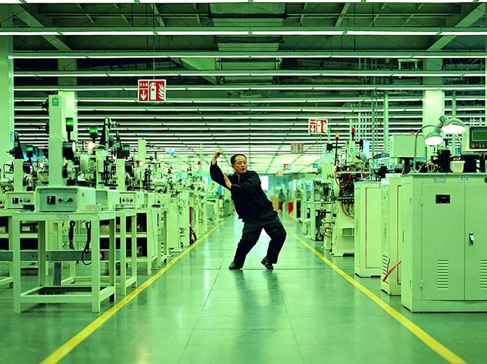 My_Future_is_Not_a_Dream Filmstill aus „Whose Utopia“ von Cao Fei aus dem Jahr 2006: Ein Mann nimmt eine tänzerische Pose in einer langen, grün beleuchteten Fabrikhalle zwischen Maschinen und Produktionsanlagen ein.